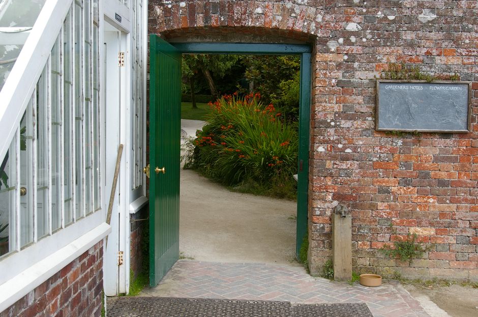 A garden entrance with a brick wall and a green-painted wooden gate, slightly open to reveal a lush garden beyond. The garden features vibrant, orange-red flowering plants with long, arching green lea