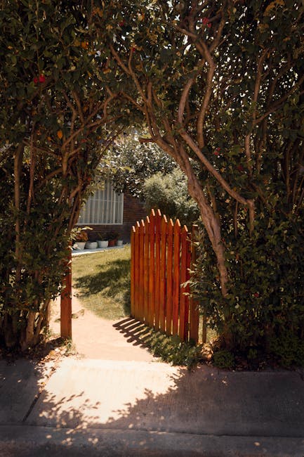 A garden entrance with a brick wall and a green-painted wooden gate, slightly open to reveal a lush garden beyond. The garden features vibrant, orange-red flowering plants with long, arching green lea